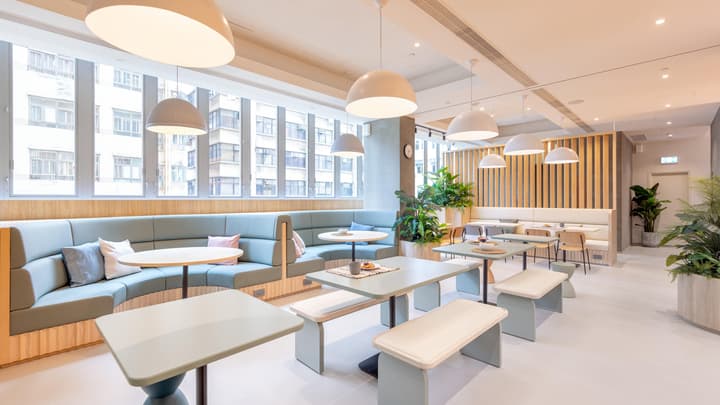 Modern communal dining and lounge area featuring long mint-green upholstered bench booths, light tables, and pendant lights, with large windows overlooking city buildings.