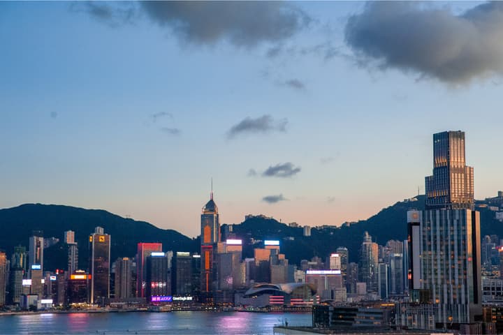 Wide shot of the dense Hong Kong skyline and harbor at dusk, showing neon and illuminated buildings against the dark mountains and a deep blue sky.