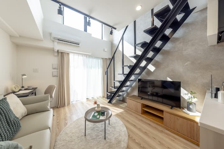 A bright, modern loft apartment living space featuring a comfortable sofa, a TV console, light wood floors, and an imposing black industrial staircase leading to the upper level.