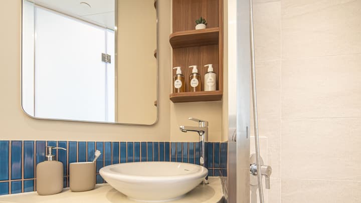 Close-up of a modern bathroom counter with a white vessel sink, chrome faucet, and wooden shelf. The wall is accented by a band of glossy, vertical blue subway tiles.