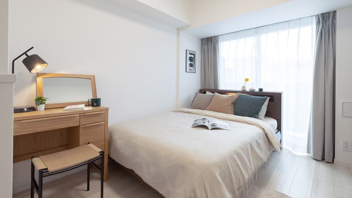 Bright, minimalist Japanese-style bedroom featuring a double bed, light wood writing desk with a lamp, and sheer curtains over a large window.
