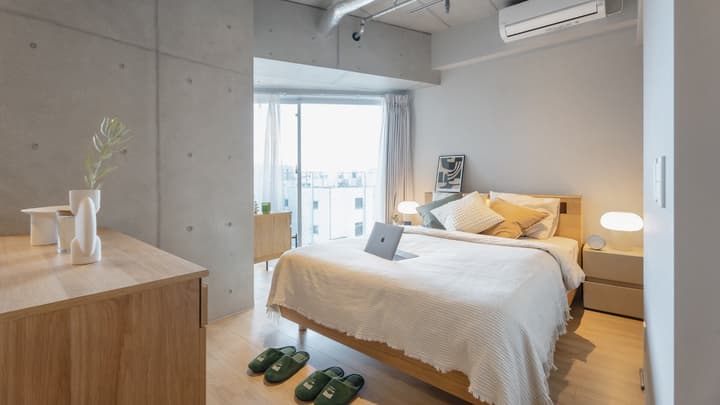 Bedroom with a light wood bed frame, white and mustard-colored bedding, and exposed concrete column and ceiling accents, with a large window and city view in the background.