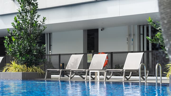 A row of four grey lounge chairs beside a sparkling dark blue swimming pool at a residential property, with lush green plants.