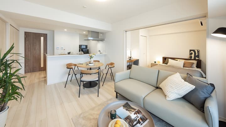 Wide-angle view of the apartment's living, dining, and kitchen area, showing a light sofa and dining table, with a sliding door leading to an adjacent private bedroom.