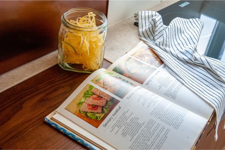 Close-up of a recipe book open to a 'Savoury Meatloaf' recipe, lying next to a jar of dried pasta and a striped kitchen towel on a wooden and stone countertop.