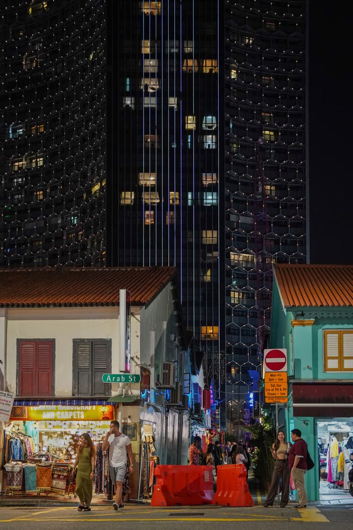 A night view of Arab Street in Singapore, showing historic, low-rise shophouses with bright storefronts contrasting sharply with a tall, modern skyscraper illuminated with vertical blue lights and tiny facade lights in the background.