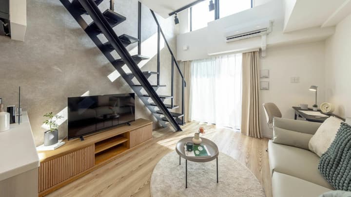 A stylish, furnished loft apartment living room with an industrial black metal staircase leading to the upper level, a media console, and large windows with sheer curtains.