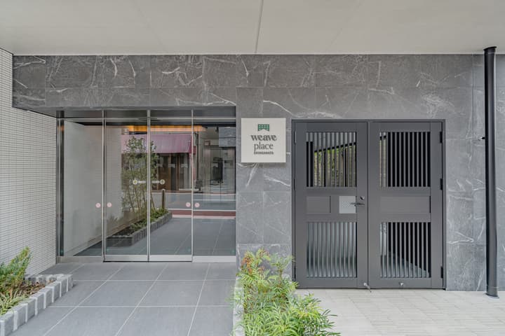 The main entrance of the apartment building, "Weave Place Shinkamata," featuring a modern design with gray marble cladding, sliding glass doors, and a decorative black metal gate.