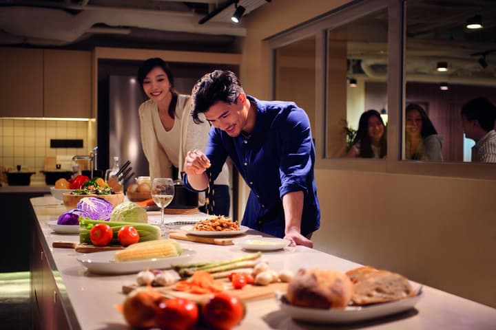 Two residents preparing food and cooking together at a large central island in the shared communal kitchen, with other residents visible through a window in the background.
