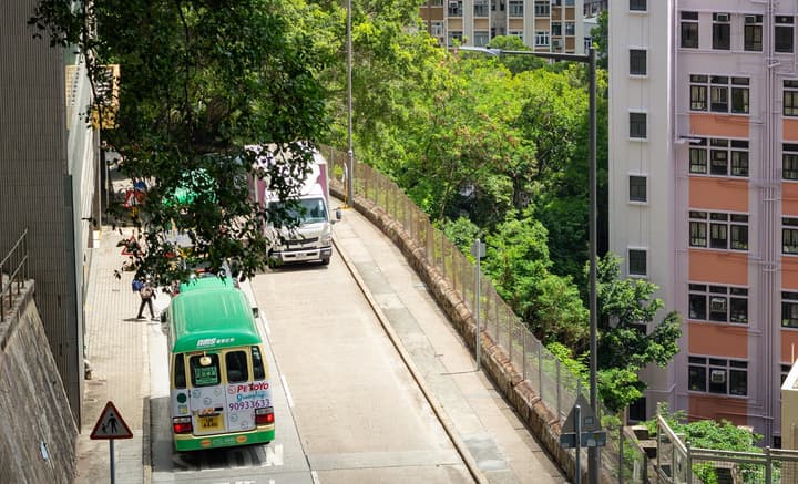 A green Hong Kong mini-bus driving up a steep, sloped road, surrounded by lush green trees and tall urban apartment buildings.