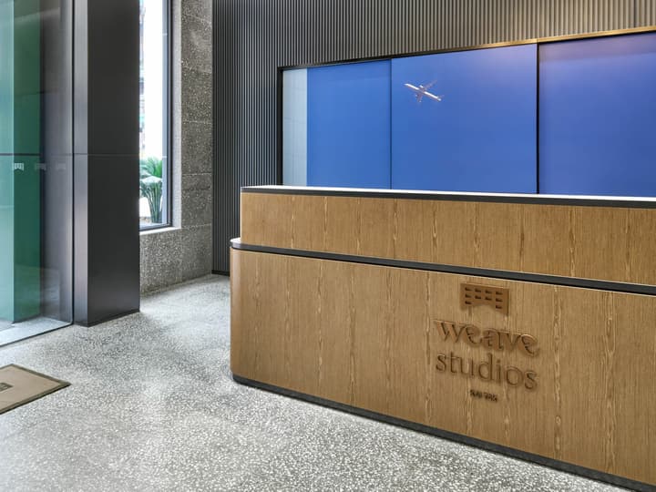 Modern coliving lobby reception desk made of blonde wood, with the "Weave Studios Kai Tak" logo, and a blue wall featuring a small airplane model.