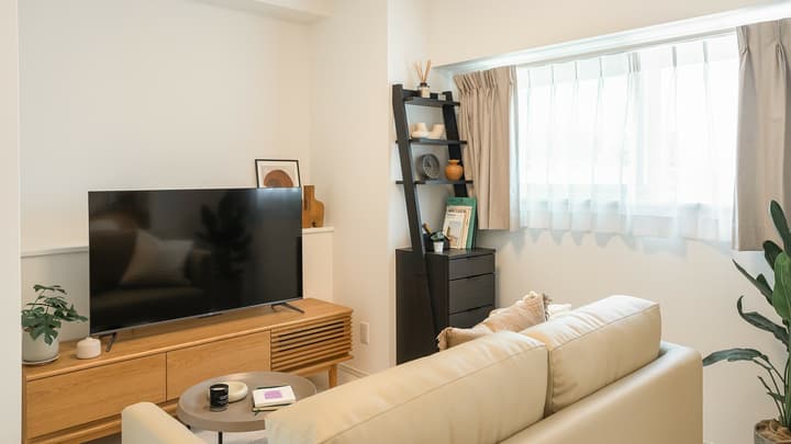 Modern living area with a light beige sofa, a low wooden entertainment center with a large TV, and a black ladder-style shelving unit next to a window.