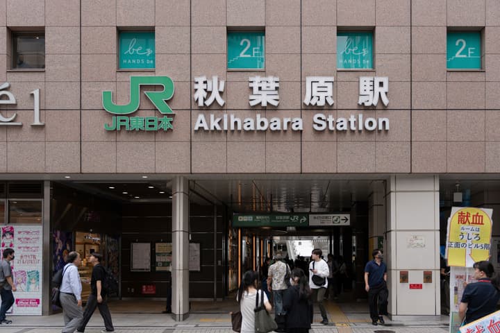 Exterior of the Akihabara Station entrance in Tokyo, showing the large JR East logo and English/Japanese station name signs above a steady flow of pedestrians.