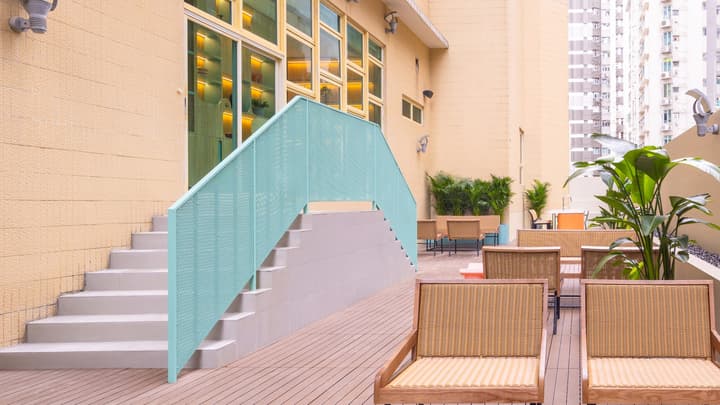 Outdoor terrace on a lower floor with wood decking, rattan-style chairs, and a staircase with a mint-green perforated metal railing leading to a doorway.