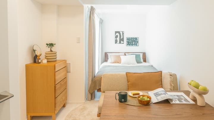 Interior view of a studio apartment showing the dining area in the foreground and the bed nook separated by a curtain in the background, with a light wood dresser and potted plant on the left.