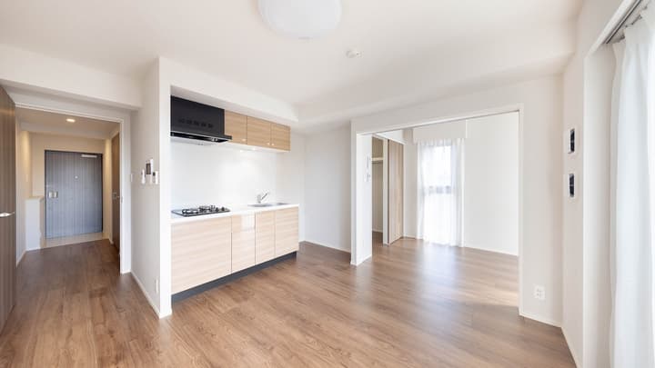 Open-concept living and kitchen area of an apartment unit with light hardwood floors, a compact kitchen counter and range hood, and sliding doors leading to a separate room.
