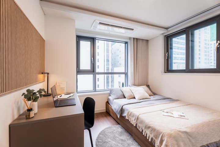 Compact bedroom featuring a single bed, a window with a city view, and a built-in desk with a laptop next to a vertical wood-slat accent panel on the wall.