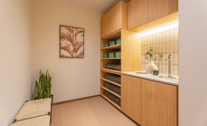 Yoga studio preparation area featuring light wood cabinets, shelving for yoga mats and blocks, and a sleek water refill station with beige tiled backsplash.