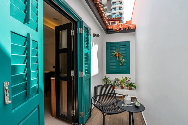 Small outdoor balcony or terrace with vibrant teal wooden shutters, white walls, a black woven chair, and a small side table next to an open door leading indoors.