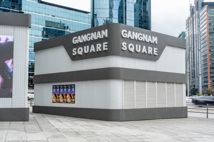 A minimalist, gray outdoor pavilion structure with large white block letters reading "GANGNAM SQUARE" on a paved sidewalk, surrounded by modern high-rise buildings.