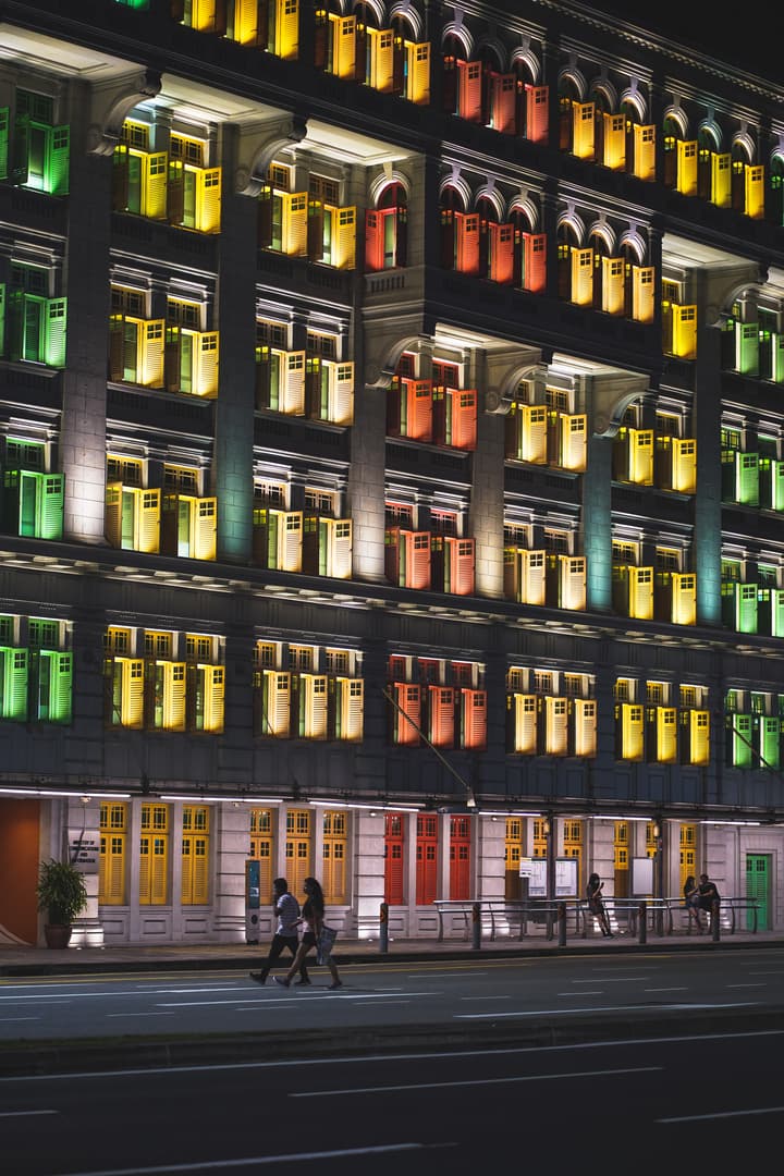 The Old Hill Street Police Station in Singapore at night, showing its symmetrical facade illuminated with windows featuring shutters painted in vibrant red, yellow, and green, with pedestrians walking in the foreground.