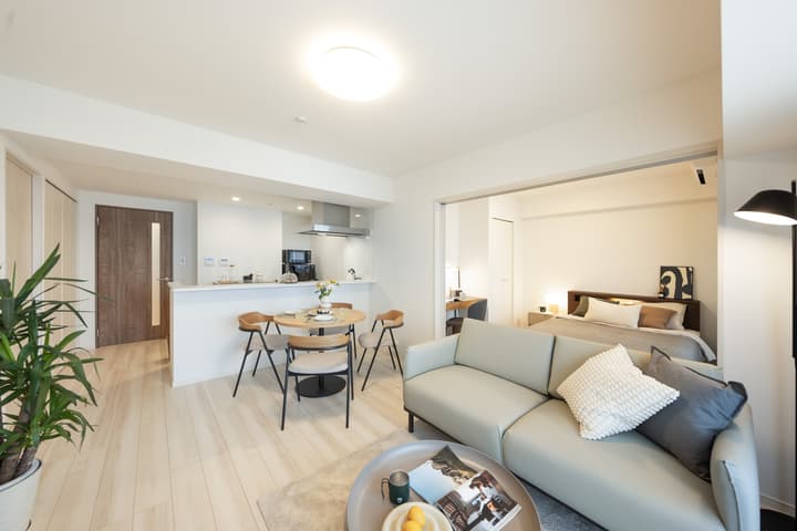 Open-concept living space with a sofa and dining table, looking toward the kitchen counter, with a wide-open doorway leading into a brightly lit bedroom.