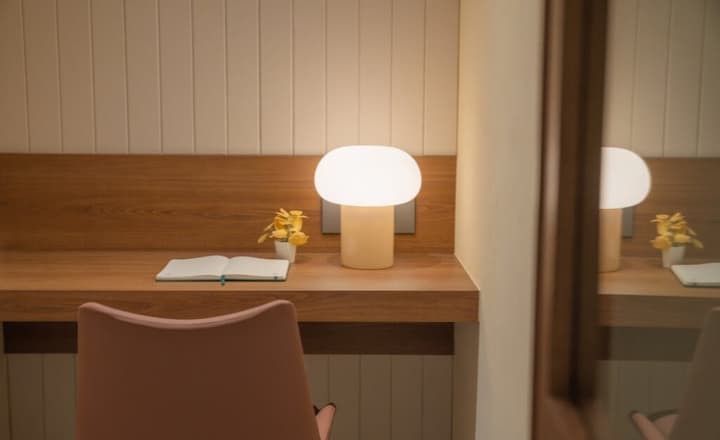 A close-up of a built-in wooden desk with an open notebook, a small yellow plant, and a modern, glowing pastel table lamp.