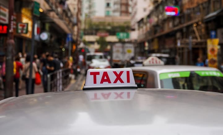 A close-up, low-angle shot of the illuminated "TAXI" sign on the roof of a vehicle. The background is a blurred, busy city street scene with people, traffic, and old buildings.