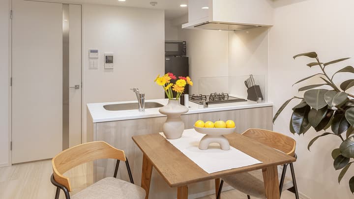 A dining area in a furnished apartment with a dark wood table set with flowers and fruit, providing a view into the compact kitchen with a cooktop and stainless steel hood.
