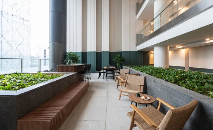 Outdoor communal terrace area with built-in wooden bench seating, natural planters, wicker chairs, and a bar area with high stools.
