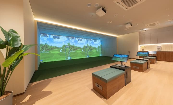 A wide indoor golf practice facility with three simultaneous simulation screens and light wood flooring.