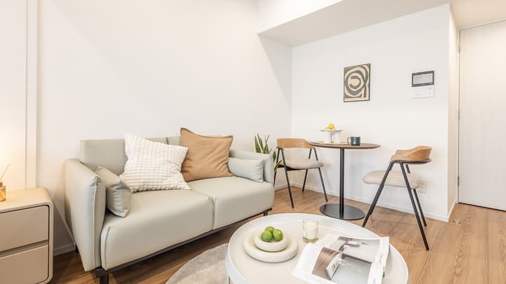 Furnished living and dining area with a light grey two-seater sofa, a small white coffee table, and a small dark wood dining table with two chairs under a modern piece of wall art.