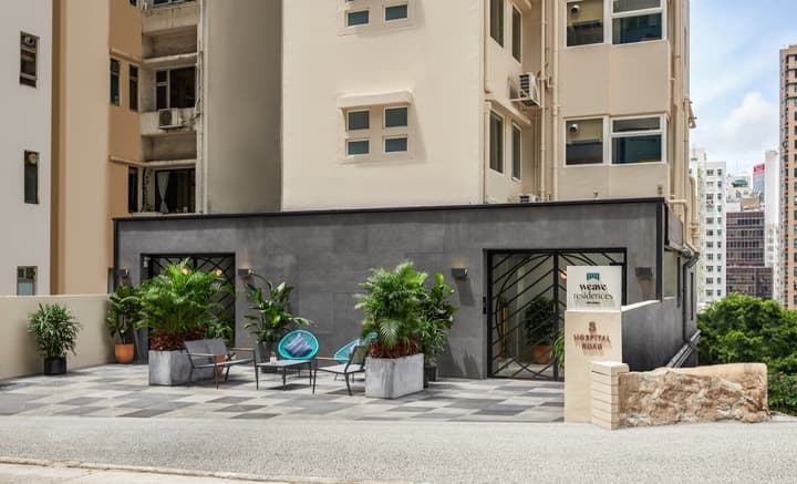 The exterior of a modern residential building featuring a sign for 'Weave Residences' and a tiled outdoor patio with planted greenery and casual seating.