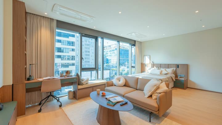 Wide interior shot of the spacious Junior Luxe Suite, featuring a queen-size bed, a tan leather two-seater sofa lounge area, a dedicated window-facing desk, and floor-to-ceiling windows with city views.