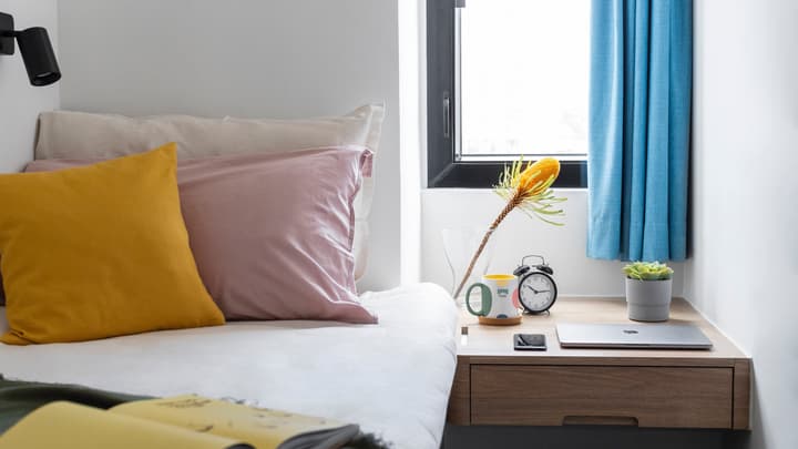 Close-up of a single bed's bedside area, featuring a small wooden nightstand with a partially opened laptop, an analog alarm clock, a yellow flowering plant in a vase, and pillows in white, soft pink, and mustard yellow.