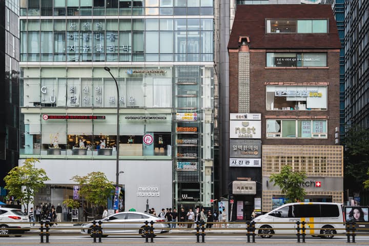 A bustling street view in Gangnam, Seoul, showing modern glass towers next to a traditional brick building, with various Korean and English shop signs and pedestrians.