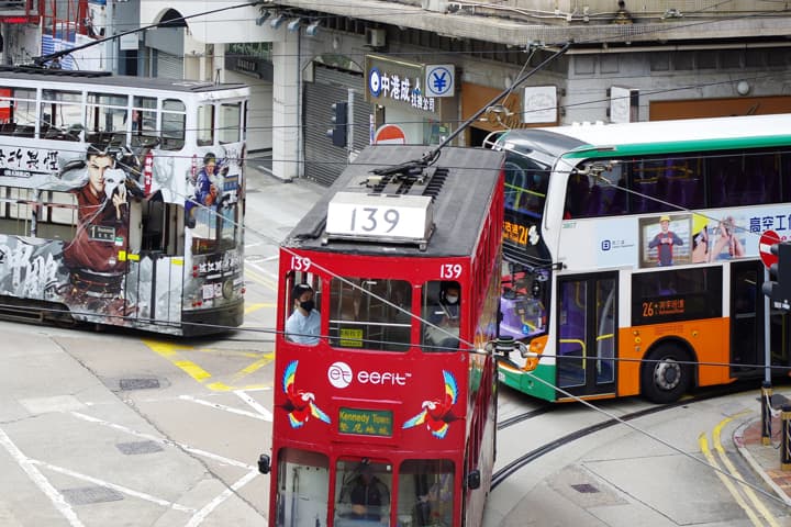 High-angle view of a busy street in Hong Kong with a red, vintage-style tram (No. 139) in the foreground, a white tram covered in large graphic advertisements on the left, and a white and green double-decker bus on the right.