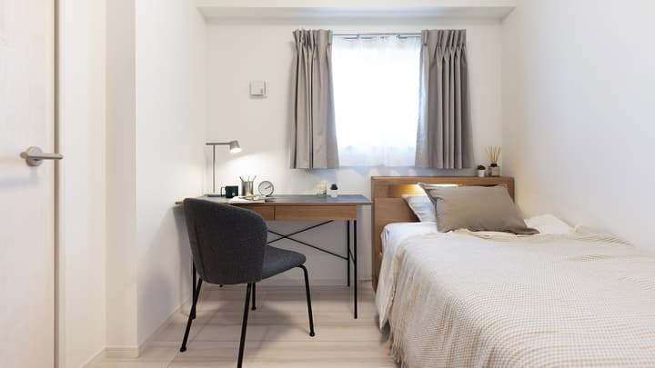 Second bedroom featuring a single bed with light wood headboard and a small study area with a wooden desk, gray chair, and desk lamp next to a curtained window.
