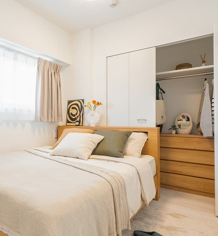 Bedroom with a wooden double bed and a sliding door closet opened to show a wooden chest of drawers and hanging clothes inside, next to a window.
