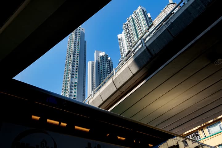 A dramatic, low-angle shot from beneath a dark concrete overpass, framing modern high-rise residential buildings against a bright blue sky.