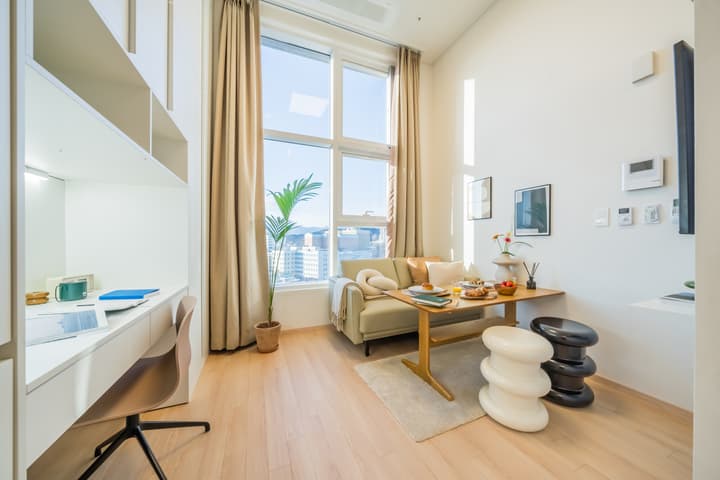 View of an apartment living area featuring a sofa, dining table, two spiral stools, and a bright built-in desk area, under tall windows with a city view.