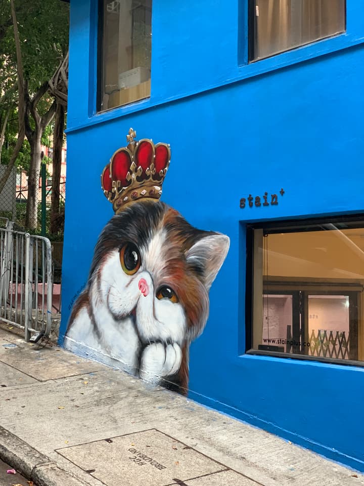 Vibrant street art mural of a large, cartoonish calico cat wearing a gold and red crown, painted on the bright blue exterior wall of a building.