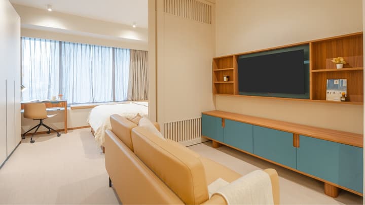 View of the junior suite showing the tan sofa and TV wall cabinet in the living area, separated by a partition from the bedroom and desk area.