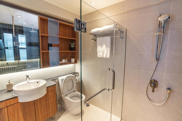 Bright, modern bathroom in a Garden Suite with a walk-in glass shower, light gray wall tiles, a wood-look vanity with a sink, and built-in wooden shelving above the toilet.
