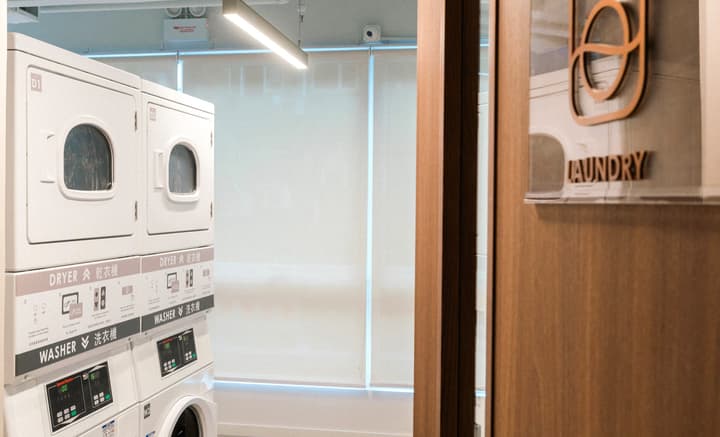 Clean, modern communal laundry room with several stacked commercial-grade washers and dryers visible, alongside a wood-framed glass door with a 'LAUNDRY' sign in a copper-colored circular graphic.