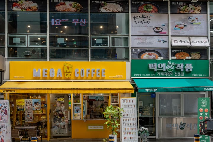 The brightly colored yellow storefront of a MEGA COFFEE shop in Korea, next to a green 'Tteok' (rice cake) shop, both with large signs and menu boards.