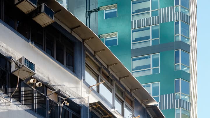 A diagonally split photograph contrasting the facade of an older building with exposed air conditioning units and awnings against a modern, sleek teal-colored skyscraper.