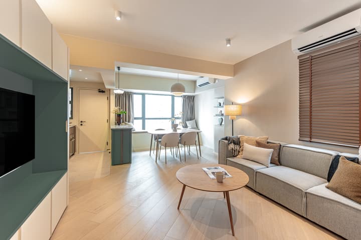 A bright, contemporary apartment living and dining area with light wood flooring, a gray modular sofa, a round coffee table, and built-in teal shelving next to the window.