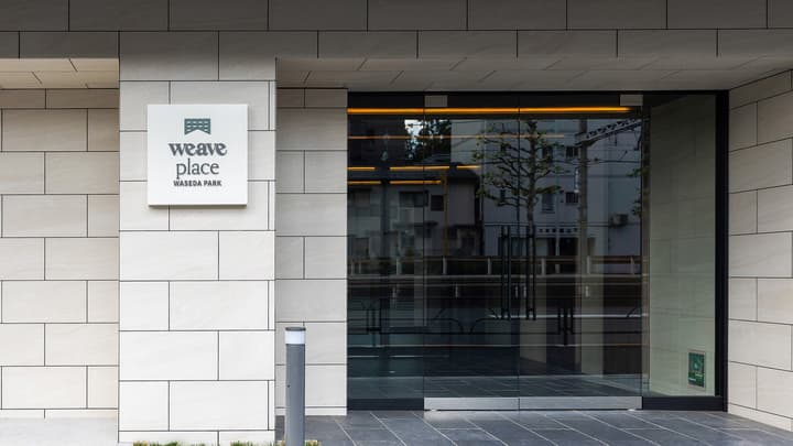 The modern, tiled main entrance of the 'Weave Place Waseda Park' residential building, featuring a sign with the logo and a clear glass facade.