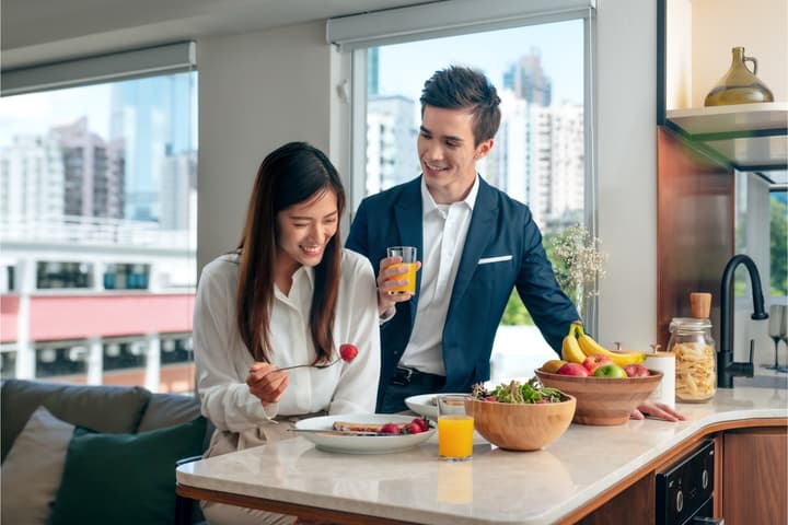 Smiling couple enjoying a healthy breakfast in a modern apartment kitchen with large windows and a bright city view in the background.